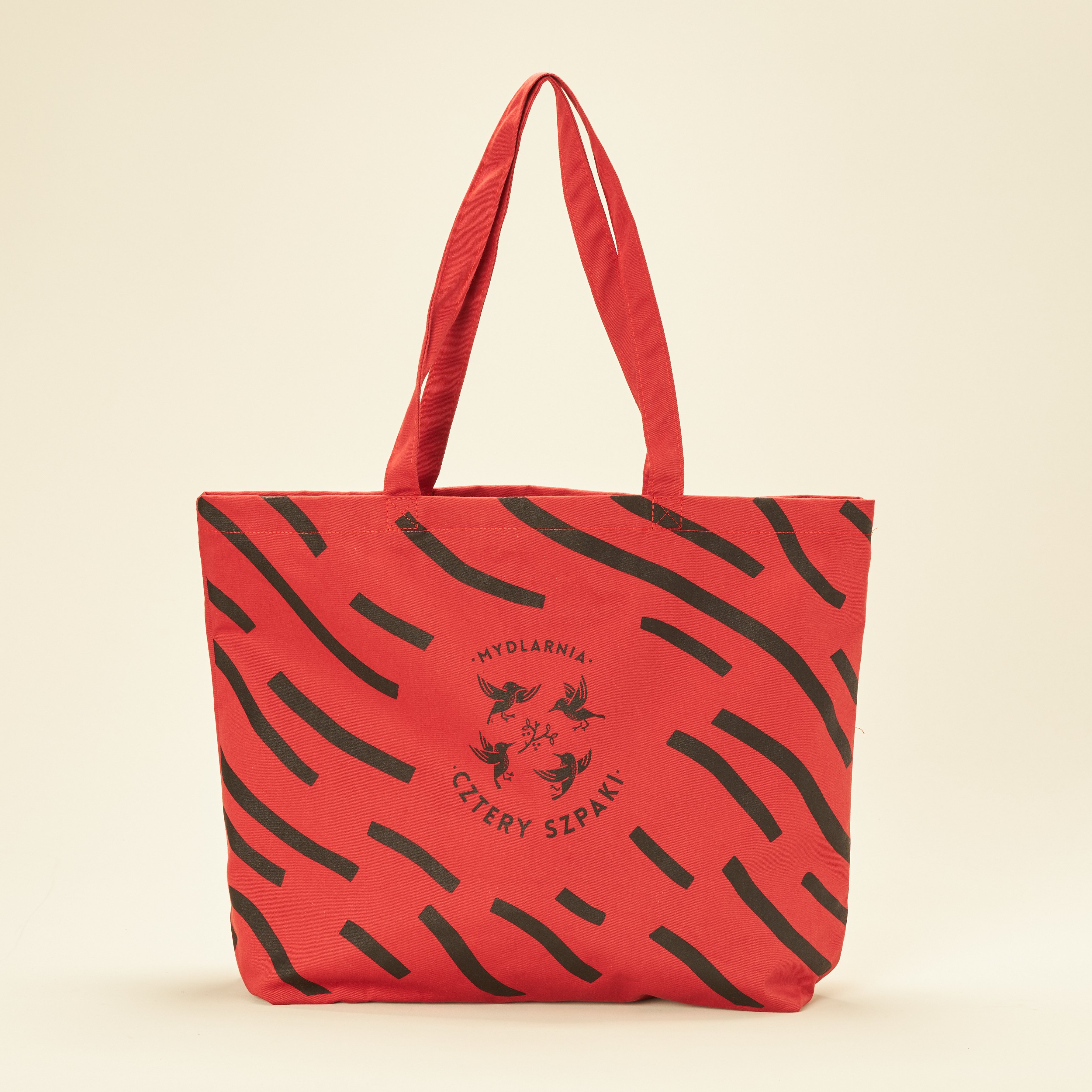 Czerwona torba z czarnymi abstrakcyjnymi paskami i okrągłym logo w środku z napisem Mydlarnia Cztery Szpaki, przedstawiającym cztery ptaki. Uchwyty są wykonane z tego samego czerwonego materiału co torba.