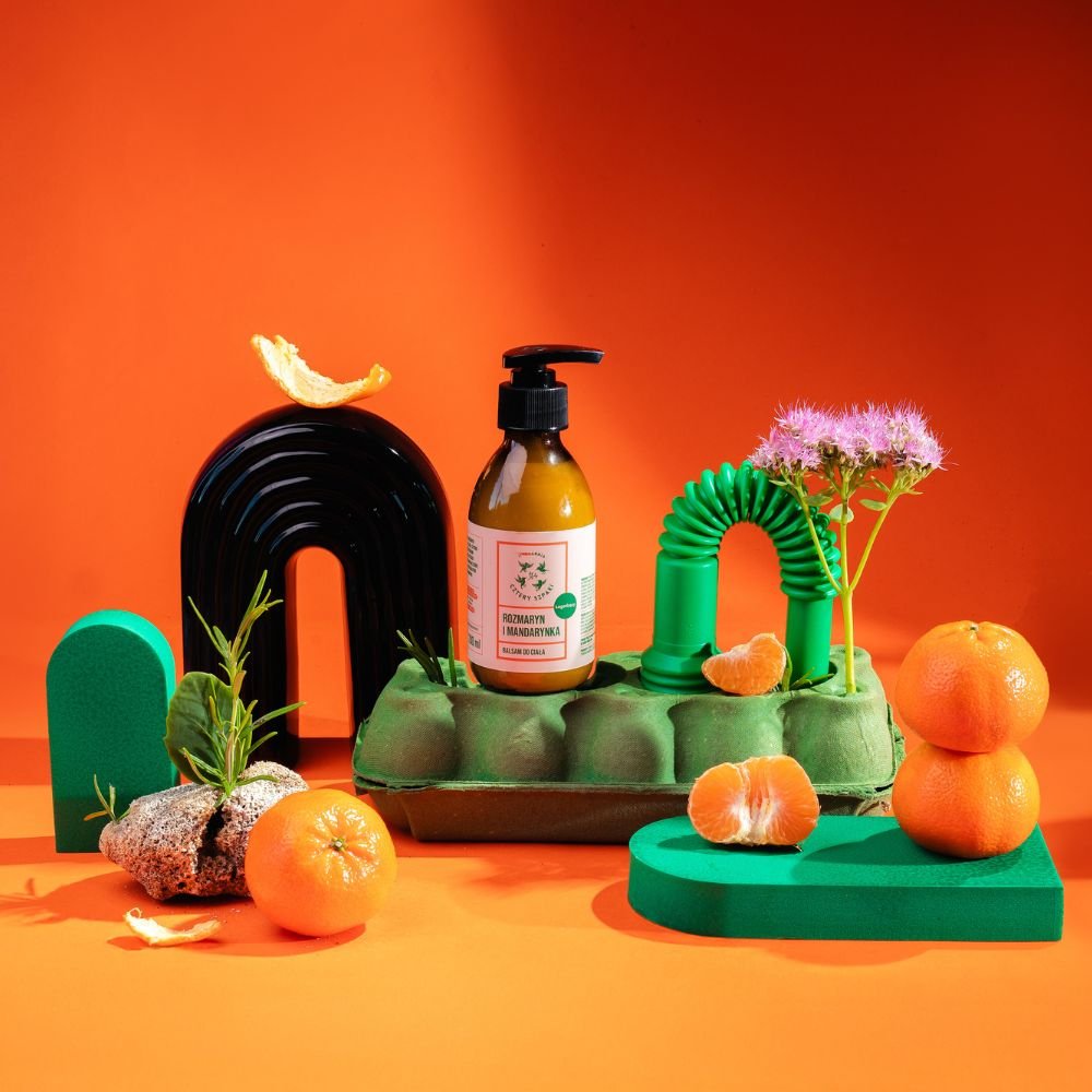 Butelka balsamu do ciała z pompką stoi na zielonym kartonie, otoczona żywymi rekwizytami na pomarańczowym tle. Świeże mandarynki, gałązka rozmarynu na kamieniu i fioletowy kwiat tworzą żywą i kolorową kompozycję.