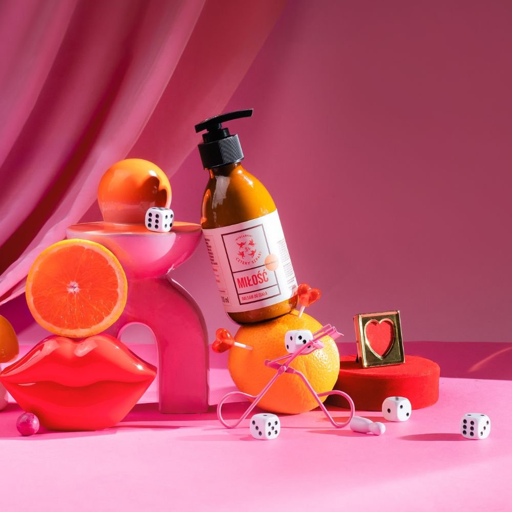 Butelka balsamu z napisem Miłość stoi na pomarańczy, otoczona kostkami do gry, różowymi kieliszkami w kształcie serca, pokrojoną pomarańczą, różowymi ustami i małym sercem w ramce, a wszystko to na różowym tle.