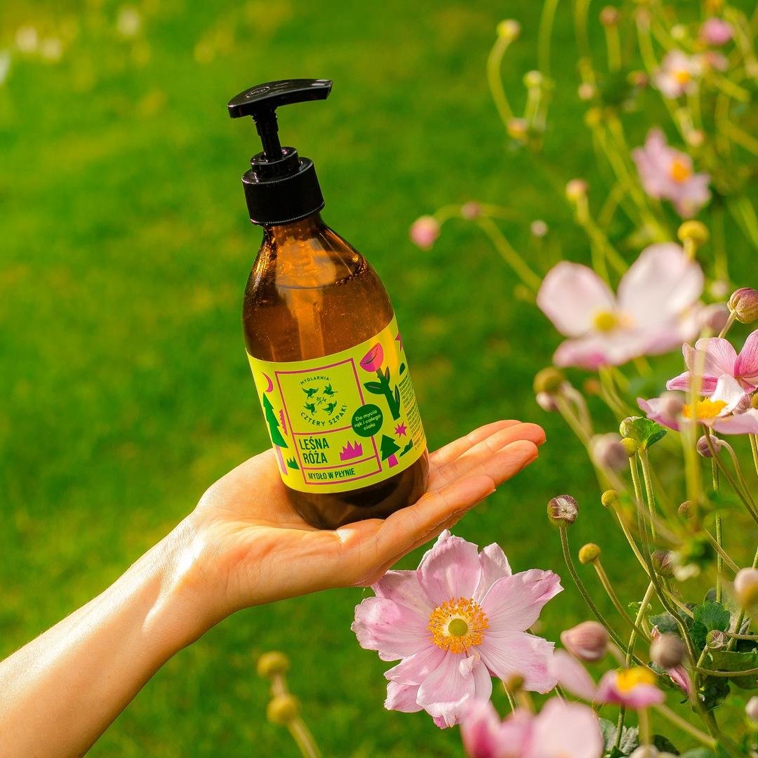 Zdjęcie mydła w płynie Leśna Róża w szklanej butelce z pompką na tle kwiatów. 