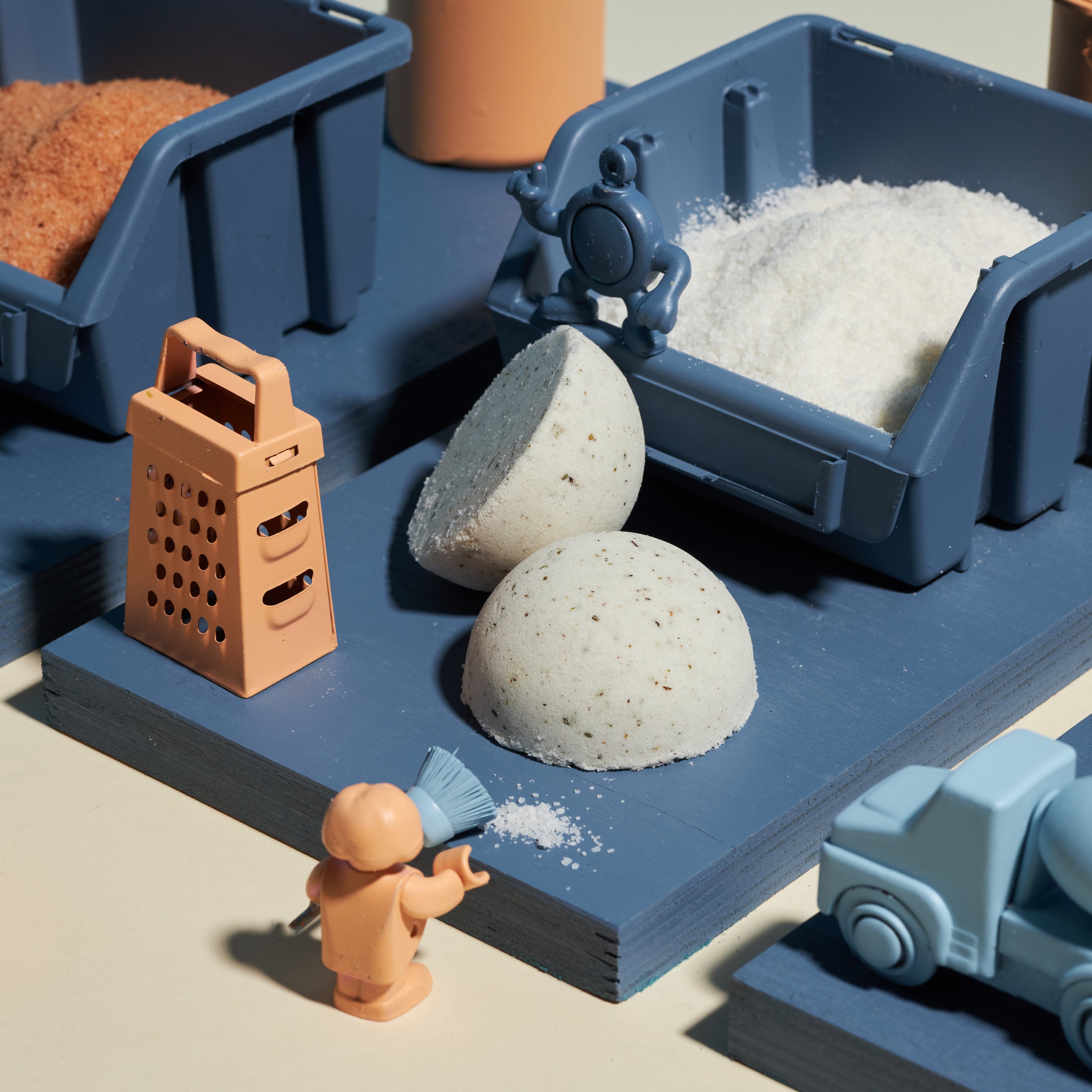 Zabawna scena zawiera miniaturowe figurki i przedmioty, w tym małą tarkę, malutką figurkę zamiatającą sól i dwie półkule do kąpieli. Ustawione są na niebieskiej platformie obok niebieskich pojemników wypełnionych granulatami i zabawkowej ciężarówki.