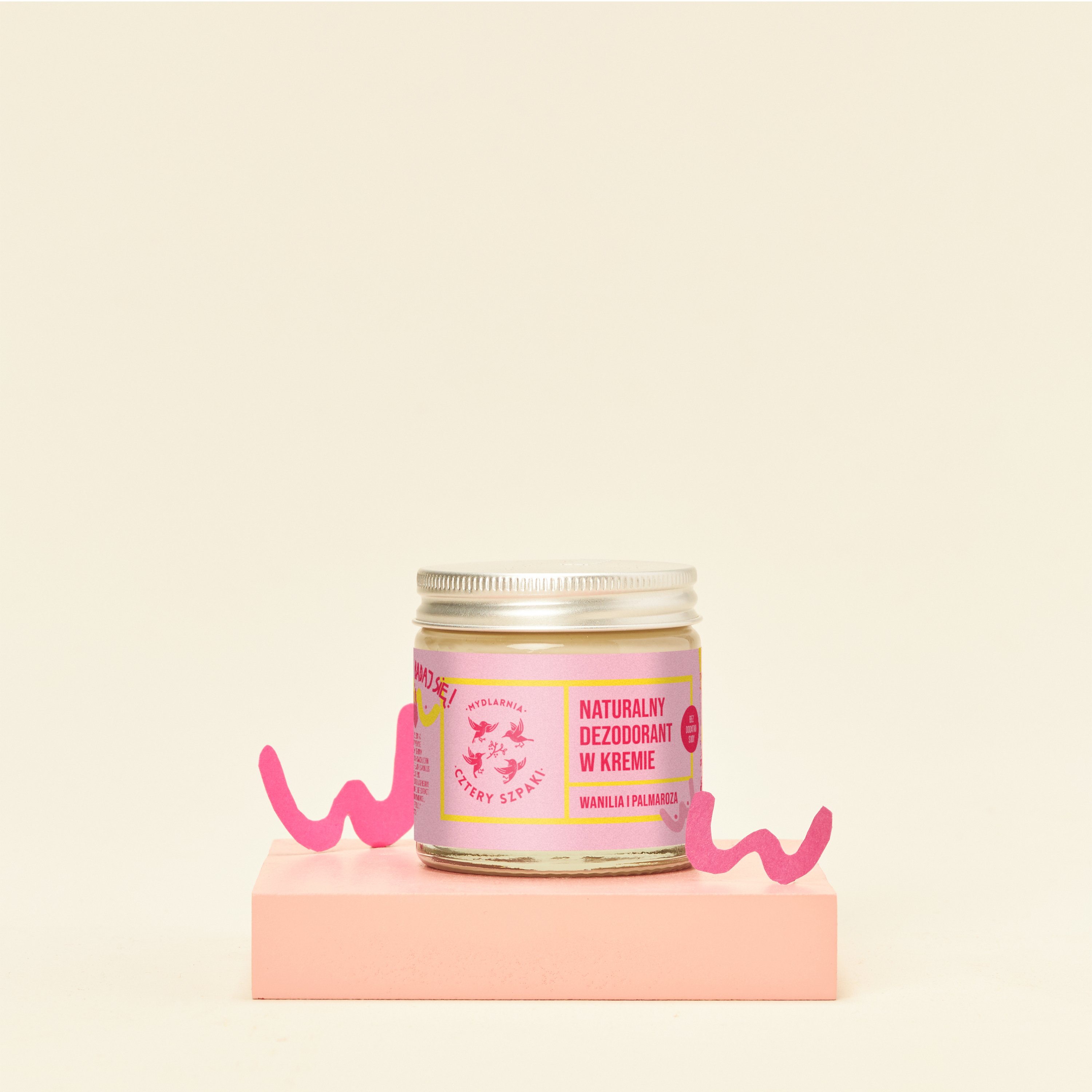 Słoiczek naturalnego dezodorantu w kremie oznakowanego w języku polskim o zapachu wanilii wyeksponowany na różowej platformie na jasnym tle.