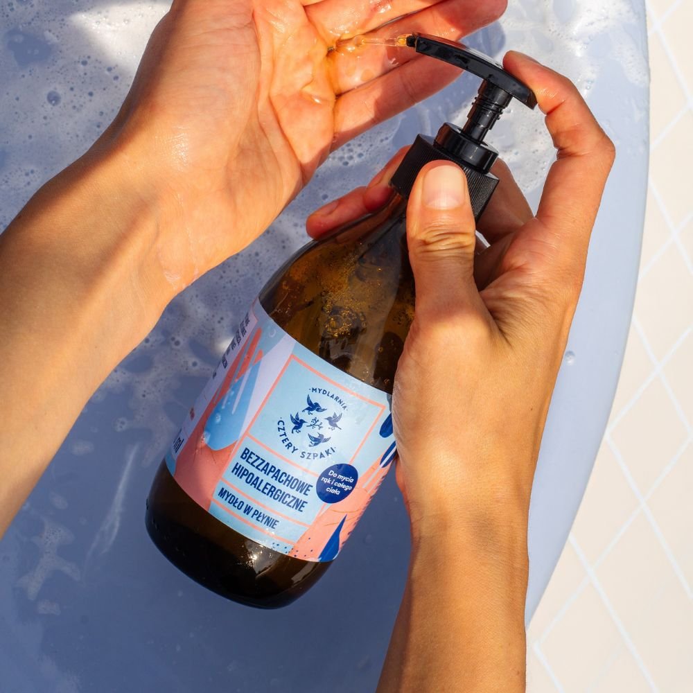 Osoba dozuje mydło w płynie z butelki z pompką do dłoni. Butelka posiada kolorową etykietę z tekstem w języku polskim, a w zestawie znajduje się wanna z wodą mydlaną.