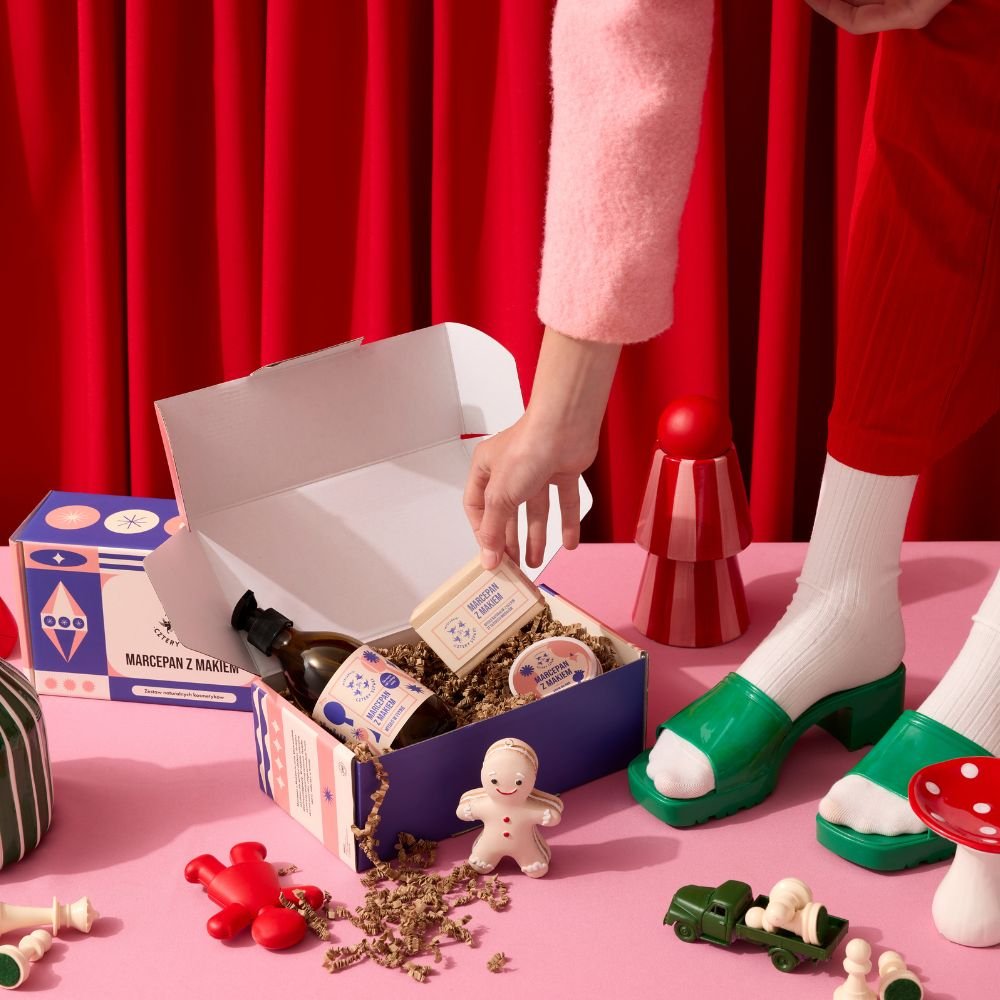 Osoba w różowym swetrze sięga do pudełka upominkowego wypełnionego produktami do pielęgnacji skóry, otoczonego zabawnymi dekoracjami, takimi jak figurka piernika i ciężarówka z zabawkami. 