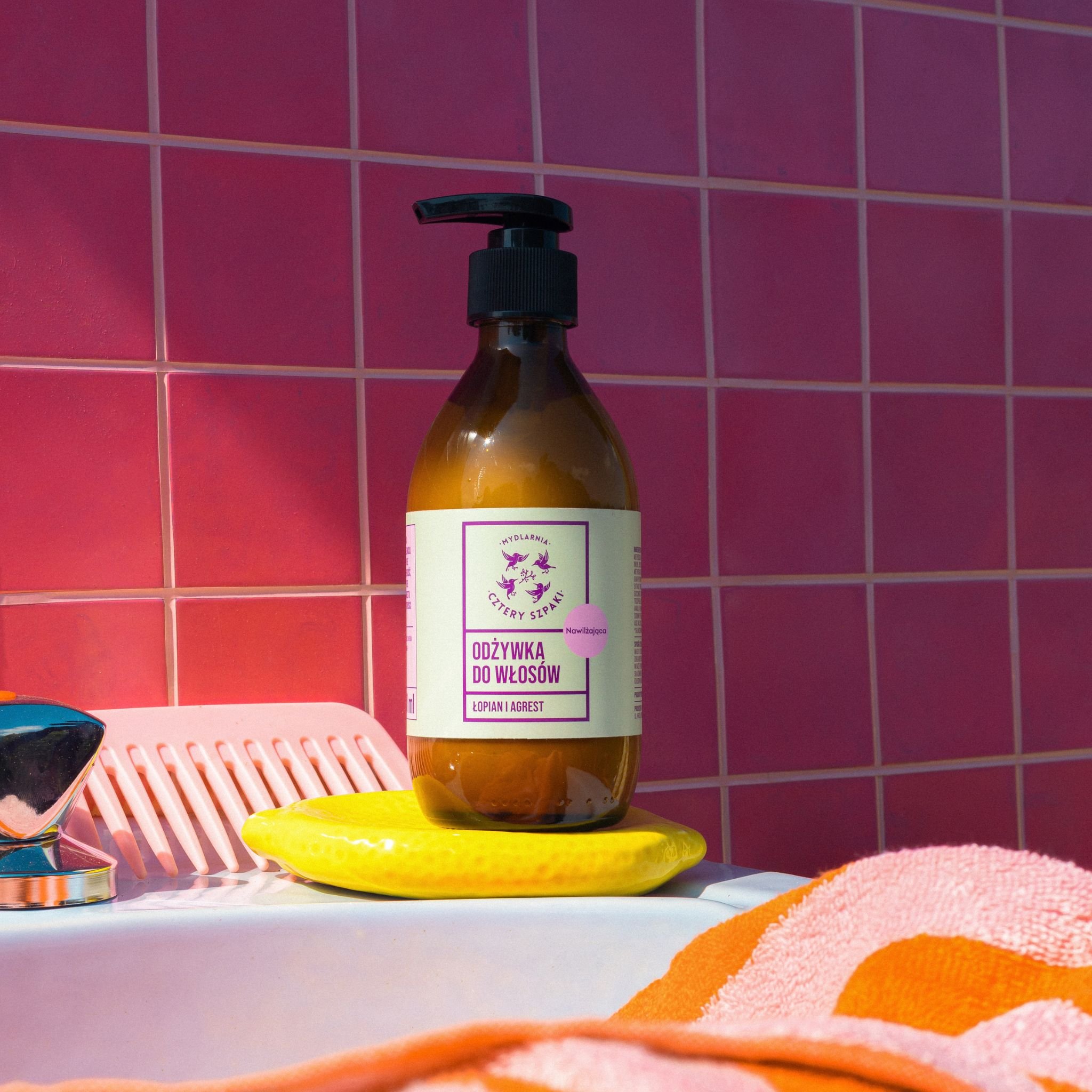 Naturalna odżywka do włosów — nawilżająca, Cztery Szpaki – Łopian i Agrest, w szklanej butelce z pompką, ustawiona w łazience z różowymi kafelkami w tle.