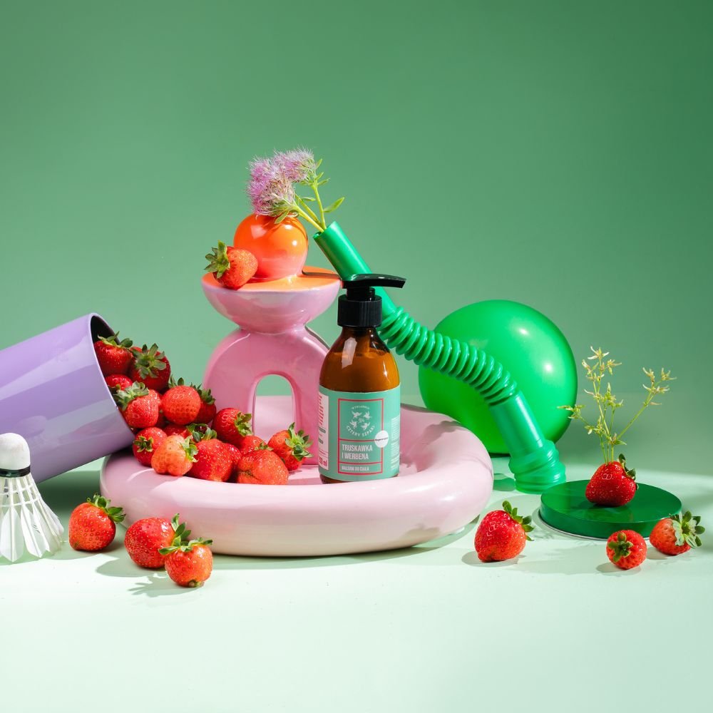 Butelka produktu do pielęgnacji skóry z pompką znajduje się wśród świeżych truskawek, zielonego balonu, pastelowych różowych i fioletowych ceramicznych przedmiotów oraz zabawnych rekwizytów, a wszystko to ułożone na zielonym tle.