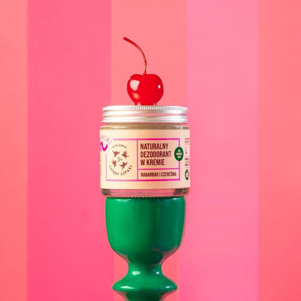 Słoik naturalnego dezodorantu w kremie stoi na zielonym cokole zwieńczonym błyszczącą czerwoną wiśnią. Na etykiecie widnieje napis „Naturalny Dezodorant w Kremie” oraz „Rabarbar i Czereśnia” na tle jaskrawego różowego paska.