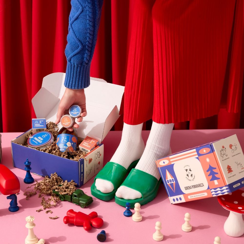 Osoba w zielonych klapkach i białych skarpetkach stoi na różowej powierzchni, otoczona kolorowymi figurami szachowymi i zabawkami. W ręku trzyma balsam i peeling i wrzuca je do pudełka z mydłem i musem Gorzka Pomarańcza. 