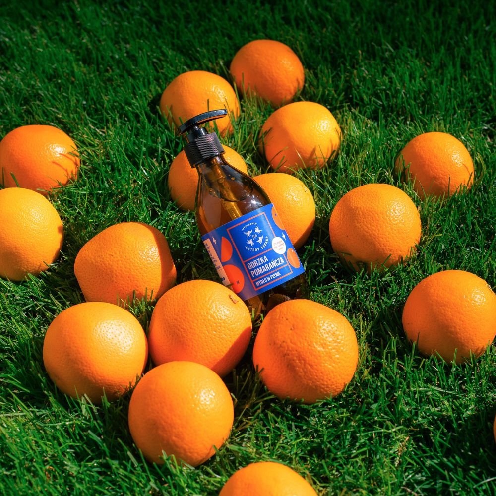 Butelka z niebieską etykietą stoi na zielonej trawie otoczona kilkoma jasnymi pomarańczami, tworząc żywy kontrast.