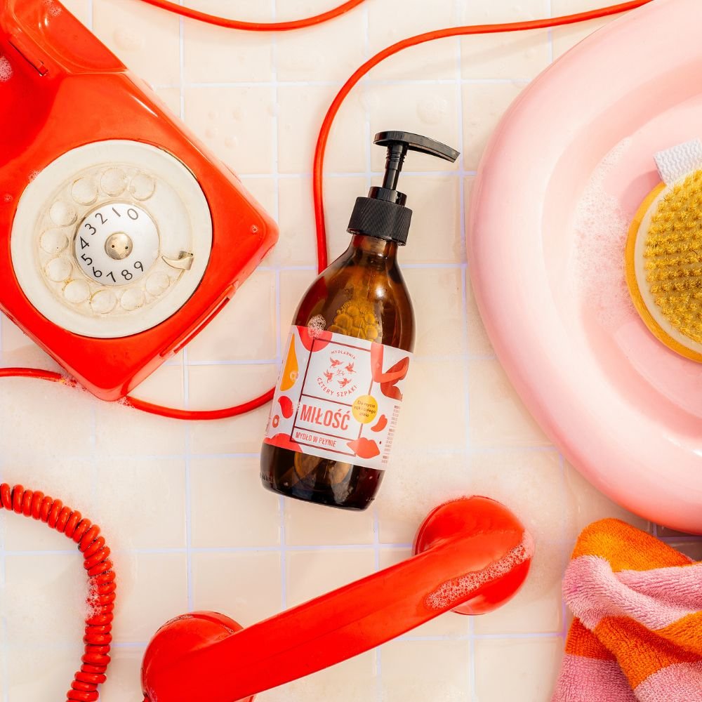Czerwony telefon obrotowy w stylu retro ze zwiniętym kablem leży na wyłożonej kafelkami powierzchni obok butelki mydła w płynie z napisem „Miłość”. Scena przedstawia różową misę wypełnioną wodą z mydłem i żółtą szczotką, z częściowo widocznym kolorowym rę