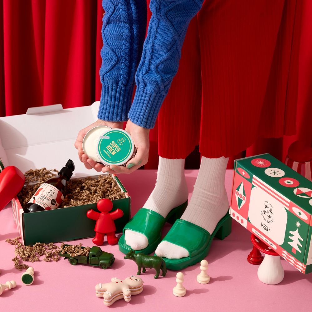 Osoba w niebieskim swetrze i czerwonej spódnicy trzyma Superkrem nad otwartym pudełkiem prezentowym wypełnionym strzępami papieru. Scena zawiera zabawne elementy, takie jak czerwona figurka, zabawkowa ciężarówka i różne małe figurki na różowej powierzchni