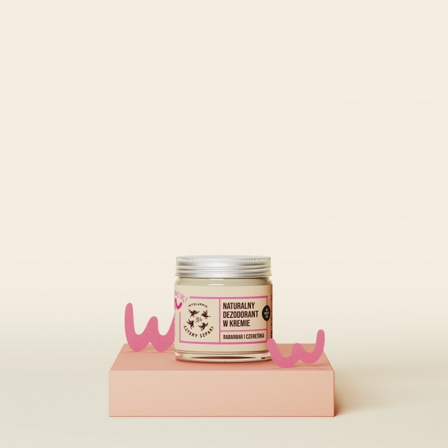 Słoiczek naturalnego dezodorantu w kremie z etykietą „Rabarbar i Czereśnia” wystawiony jest na platformie w kolorze brzoskwiniowym, z różowymi elementami dekoracyjnymi po obu stronach, na delikatnym beżowym tle.